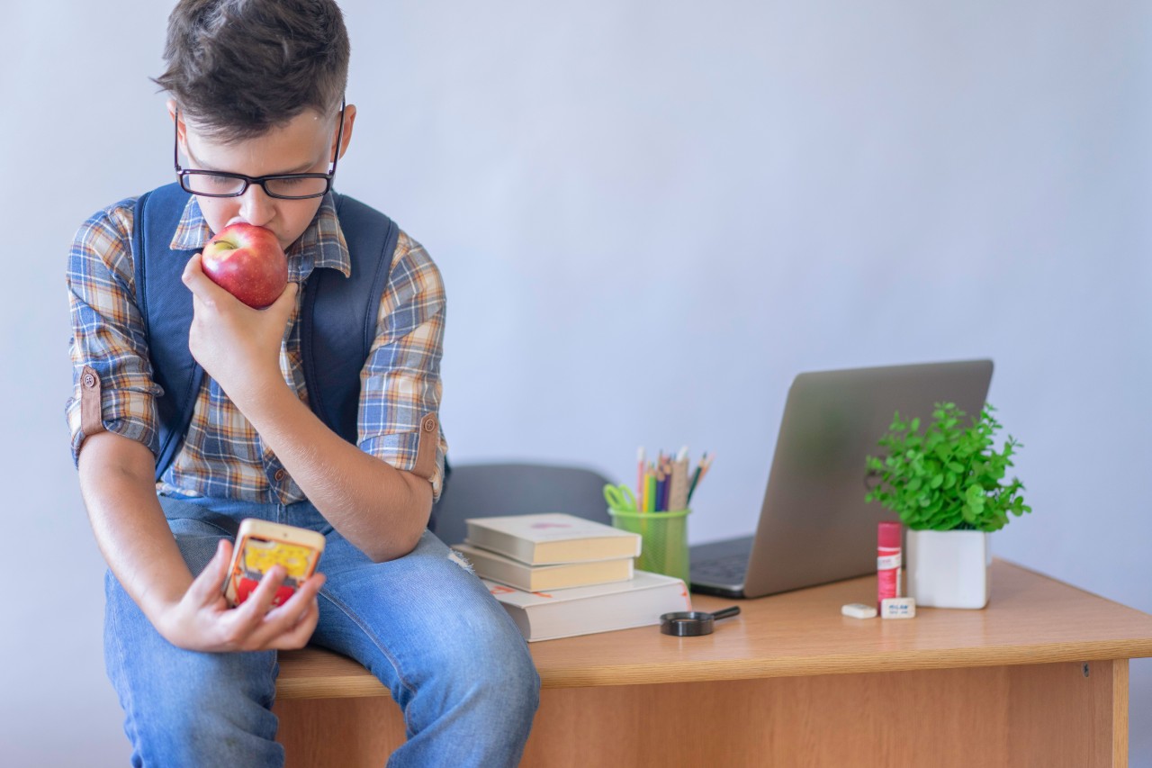 A Schoolboy with a Phone in his Hand Eats an Apple