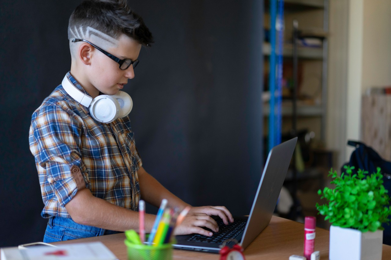 A Schoolboy in Glasses with Headphones Works on a Laptop
